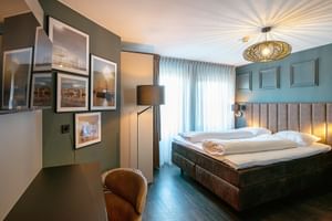 Modernes Hotelzimmer mit türkisfarbenen Wänden, beigem Polsterbett, dekorativer Deckenleuchte, gerahmten Fotos, Stehlampe und braunem Sessel.