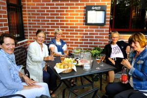 Fünf Frauen sitzen an einem Tisch im Freien mit Getränken und Snacks vor einer roten Backsteinmauer und genießen einen entspannten Moment.