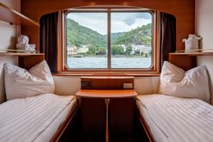 Zweibettkabine auf MS Olympia mit zwei Betten, Holzmöbeln und großem Fenster mit Blick auf Flusslandschaft mit grünen Hügeln und Ufergebäuden.