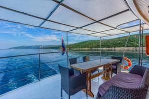 Außenbereich des Schiffes Linda mit Esstisch, Korbstühlen und Glasgeländer mit Blick auf blaues Wasser und bewaldete Küste.