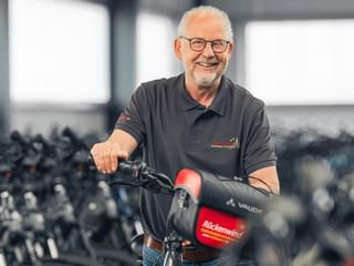 Uwer Hanusch mit Brille und grauem Poloshirt, lächelnd am Fahrradlenker in einer Werkstatt mit vielen Fahrrädern im Hintergrund.