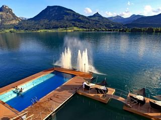 Schwimmende Holzterrasse mit blauem Pool auf dem Wolfgangsee mit Wasserfontänen und Bergkulisse in St. Wolfgang, Österreich.