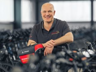 Knut Wulfestieg in dark polo shirt leaning on bicycle handlebar in workshop with rows of bikes in background.