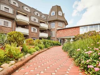 Backsteinhotel mit Turm und Balkonen in Varel. Gepflasterter Weg führt durch Garten mit bunten Blumen und grünen Pflanzen.
