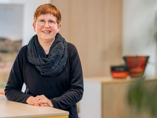 Meike Engling with short red hair and glasses, wearing a black top and patterned scarf, smiling at a desk in a bright office.