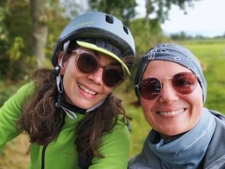 Zwei lächelnde Frauen mit Helmen und Sonnenbrillen machen ein Selfie im ländlichen Ostfriesland. Grüne Felder und Bäume im Hintergrund.