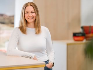 Sabrina Broziat with long blonde hair and white long-sleeved shirt, smiling in a modern office with colorful bowls and window view.