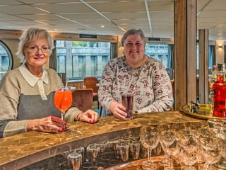 Zwei Frauen stehen an einer Marmorbar mit Getränken auf der MS Princess. Moderne Lounge mit Fenstern und Sitzgelegenheiten im Hintergrund.