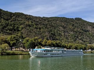 MS Switzerland Flusskreuzfahrtschiff auf ruhigem Wasser mit bewaldeten Bergen und kleinem Dorf im Hintergrund unter blauem Himmel.