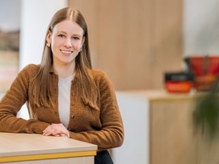 Portrait of Greta Martens with long brown hair, wearing a brown cardigan and white top, smiling at a desk in a bright office.