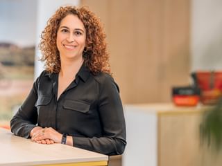 Irene Godoy Marin with curly brown hair, wearing a black shirt and watch, smiling while standing at a white desk in a modern office.