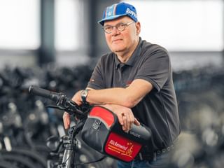 Robert Goldberg mit Brille und blauer Kappe, lehnt an Fahrrad mit roter Tasche im Fitnesscenter mit Heimtrainern im Hintergrund.