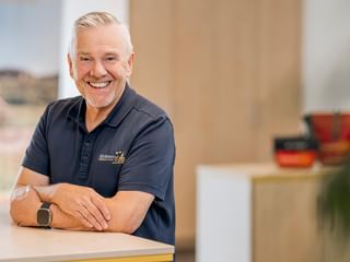 Uwe Klemme with gray hair wearing a dark polo shirt, smiling at a counter. He wears a smartwatch and stands in a bright office space.