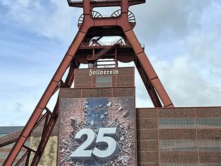 Roter Förderturm der Zeche Zollverein mit Backsteingebäude und '25'-Banner. Die ikonische Industriestruktur erhebt sich vor blauem Himmel.