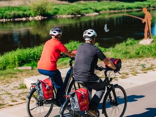 Two cyclists with helmets and touring bikes with red panniers sit on cobblestones by the Weser River. A person fishes in the background.