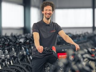 Basti Stüdemann in schwarzem Poloshirt steht mit Fahrrad in großer Lagerhalle voller Fahrradreihen.
