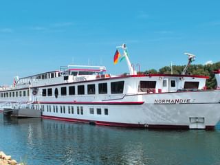 Weißes Flusskreuzfahrtschiff MS Normandie mit rotem Zierstreifen und deutscher Flagge auf ruhigem Wasser, grünes Ufer im Hintergrund.