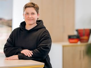 Romy Kellert with short brown hair wearing a black hoodie, standing at a light wooden table in a bright modern office space.