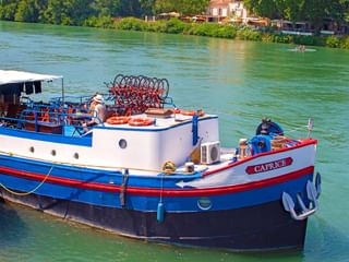 Weiß-blauer Schaufelraddampfer MS Caprice mit rotem Schaufelrad am grünen Flusswasser vertäut, mit üppiger Vegetation und Gebäuden am Ufer.