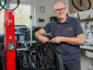 Herms Preuss with glasses and gray polo shirt smiling in a bicycle workshop, working on a bike with red repair stand and tools visible.