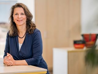 Heike Salver with shoulder-length brown hair, wearing a navy blue blouse and necklace, sitting at a light-colored desk in a bright office.