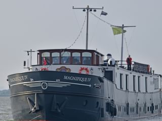 MS Magnifique IV Flusskreuzfahrtschiff auf offenem Wasser. Das Schiff hat einen dunklen Rumpf und weißen Aufbau mit großen Fenstern.
