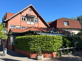 Rotes Backsteingebäude Hotel Central in Zeven mit orangefarbenem Ziegeldach und grüner Hecke. Klarer blauer Himmel und weißes Nebengebäude.