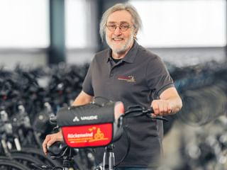 Hans Wüst mit Brille und grauen Haaren, trägt ein dunkles Poloshirt und steht mit einem Fahrrad in einer Werkstatt voller Räder.