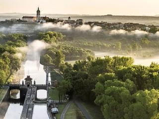 Luftaufnahme von Mělník mit Kirchturm über Morgennebel. Schleuse an der Elbe im Vordergrund, umgeben von grünen Wäldern.