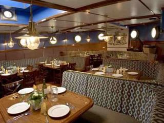 Innenraum des Mare fan Fryslan Restaurants mit Holztischen, gemusterten Banksitzen, Messingpendelleuchten und nautischem Dekor.