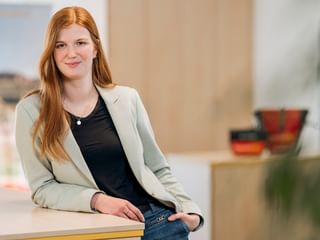 Phyllis Wiechmann with long red hair wearing a beige blazer and black top, standing in a modern office with blurred background.