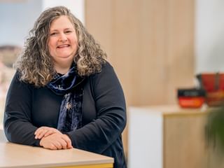 Britta Krefeldt with shoulder-length curly gray hair, wearing a dark blazer and patterned scarf, smiling in a bright office setting.