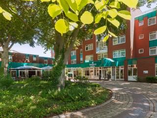 Außenansicht des Upstalsboom Hotels Emden mit roter Backsteinfassade und grünen Akzenten. Gepflasterter Innenhof mit alten Bäumen und Terrasse.