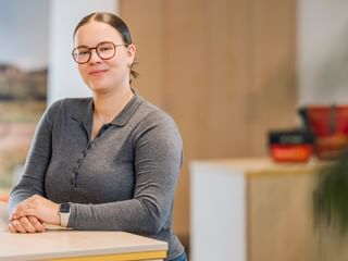Pia Wilken with glasses and hair tied back, wearing a gray long-sleeved shirt, standing at a wooden counter in a bright office space.