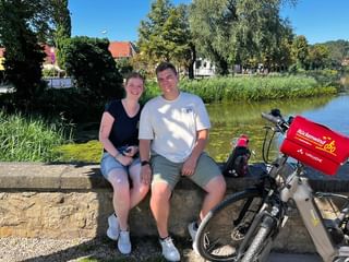 Junges Paar sitzt auf Steinmauer am See mit zwei Tourenrädern. Frau in schwarzem Oberteil, Mann in weißem Hemd. Rote Fahrradtaschen sichtbar, Häuser und Bäume im Hintergrund.