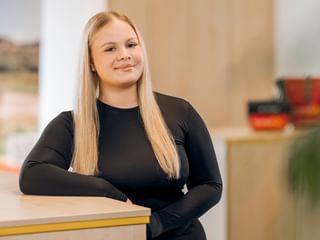 Cassandra Morla Silva with long blonde hair wearing a black long-sleeved top, standing at a reception desk in a bright office space.