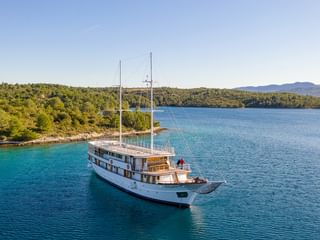 Weiße Motoryacht MY Magellan mit zwei Masten fährt durch türkisfarbenes Wasser entlang der bewaldeten kroatischen Küste unter blauem Himmel.
