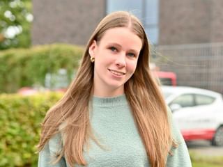 Porträt von Greta Martens mit langen blonden Haaren in einem mintgrünen Pullover, lächelt in die Kamera mit Backsteingebäude und Autos im Hintergrund.