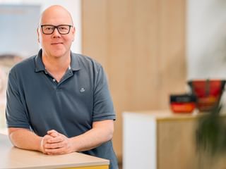 Markus Hüneke wearing black-framed glasses and a gray polo shirt, sitting at a light wooden counter in a bright office space.