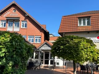 Hotel Central in Zeven mit roter Backsteinfassade und Außenterrasse. Zwei Gebäude flankieren einen Innenhof mit grünen Bäumen und Sitzbereich.