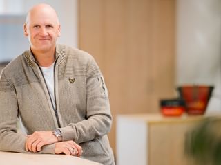 Andreas Bunge with a friendly smile, wearing a beige knit cardigan over a white shirt, standing in a modern office space.