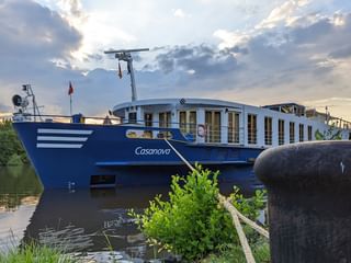 Blau-weißes Flusskreuzfahrtschiff MS Casanova liegt an einem Flussufer unter bewölktem Himmel. Grüne Vegetation und ein Anlegepfosten im Vordergrund.
