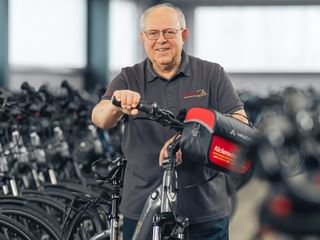 Janos Raduly mit Brille und grauem Poloshirt steht mit einem Fahrrad mit roter Tasche in einer Lagerhalle voller Fahrradreihen.