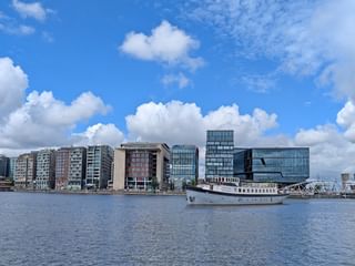 Weißes Passagierschiff Floor van Aemstel am modernen Ufer mit zeitgenössischen Glas- und Betongebäuden unter blauem bewölktem Himmel.