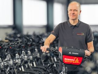 Michael Schubert in grauem Poloshirt steht mit Fahrrad vor Reihen schwarzer Fahrräder in einer hellen Lagerhalle.