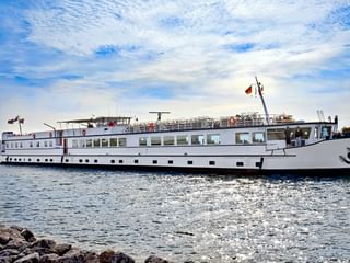 Weißes Passagierschiff DE Holland fährt auf ruhigem Wasser unter bewölktem blauen Himmel, mit felsiger Küste im Vordergrund.