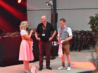 Frau im rosa Dirndl interviewt zwei Männer auf der Bühne bei Jubiläumsfeier, mit Fahrrädern und roter Beleuchtung im Hintergrund.
