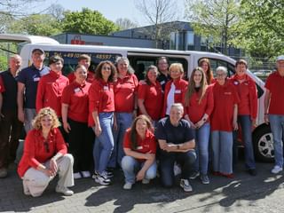 Großes Team-Gruppenfoto mit Personen in roten Shirts, die vor einem weißen Transporter stehen und knien, draußen an einem sonnigen Tag.
