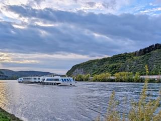 Weißes Flusskreuzfahrtschiff SE Manon auf dem Rhein bei Andernach, mit grünen bewaldeten Hügeln auf beiden Seiten unter bewölktem Abendhimmel.