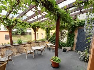 Überdachte Terrasse mit weinbewachsener Pergola in der Schlossschänke Bautzen. Steinmauern, Bistrotische und Stühle auf grauem Boden.